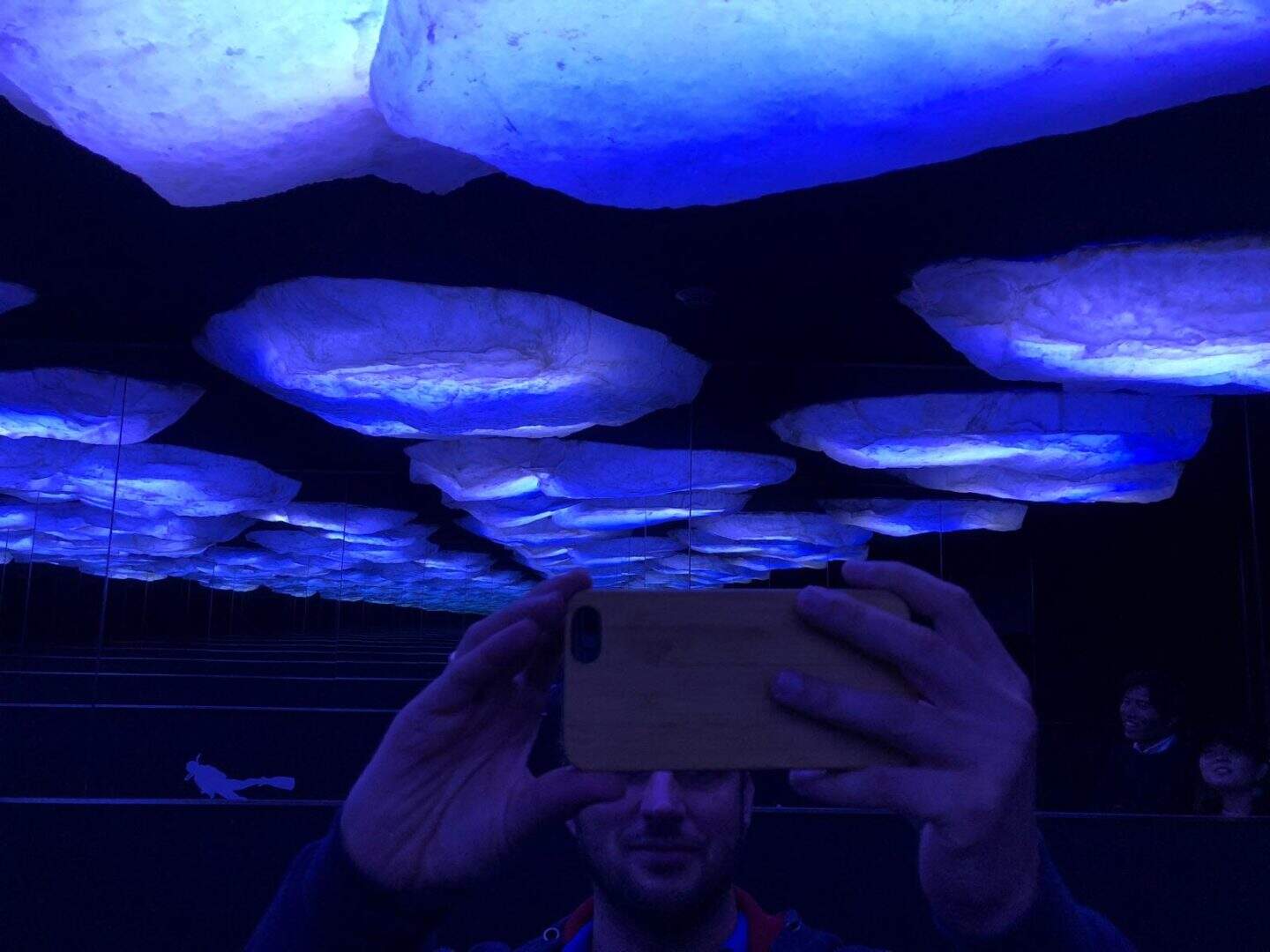 Imagem de uma pessoa tirando uma selfie em um ambiente com iluminação azul no teto, que parece uma instalação artística ou uma caverna artificial com iluminação LED azul, criando um efeito visual impr.