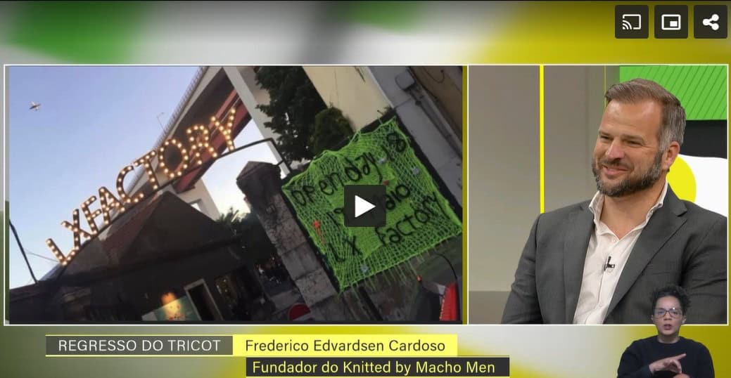 Imagem de Frederico Cardoso, fundador do Knitted by Macho Men, durante uma entrevista na RTP, com um painel de fundo e uma placa de protesto visível.