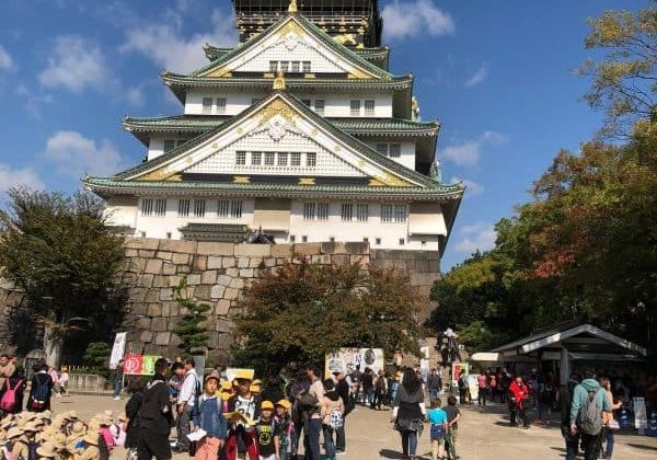 Imagem de uma torre de castelo tradicional japonesa com múltiplos andares e telhados curvos, cercada por árvores e um grupo de visitantes explorando o local sob céu azul claro.
