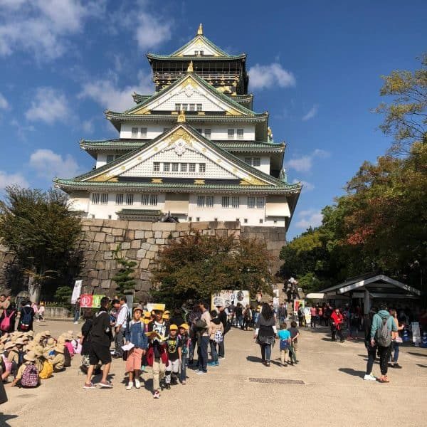 Imagem de uma torre de castelo tradicional japonesa com múltiplos andares e telhados curvos, cercada por árvores e um grupo de visitantes explorando o local sob céu azul claro.