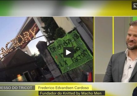 Imagem de Frederico Cardoso, fundador do Knitted by Macho Men, durante uma entrevista na RTP, com um painel de fundo e uma placa de protesto visível.
