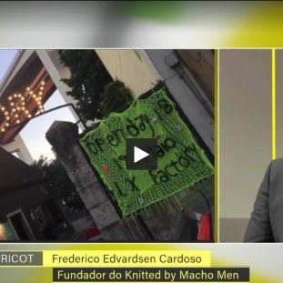 Imagem de Frederico Cardoso, fundador do Knitted by Macho Men, durante uma entrevista na RTP, com um painel de fundo e uma placa de protesto visível.